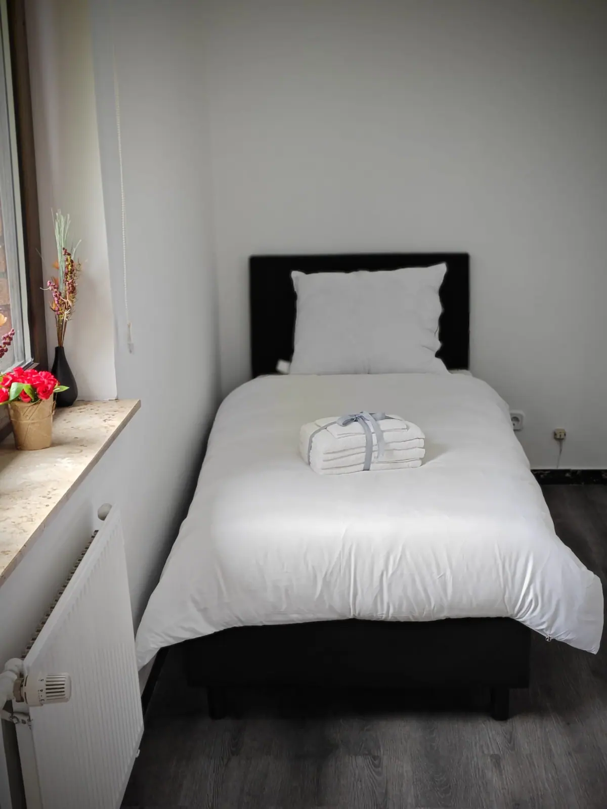 Einzelbett vor weißer Wand mit Handtuchstapel und Tageslicht vom Fenster